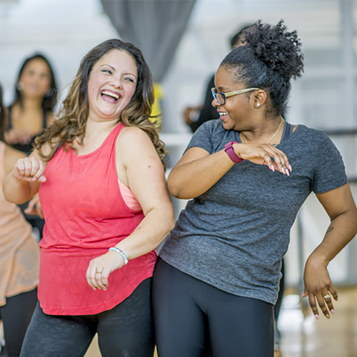 Two women laughing and dancing in a group fitness class. 