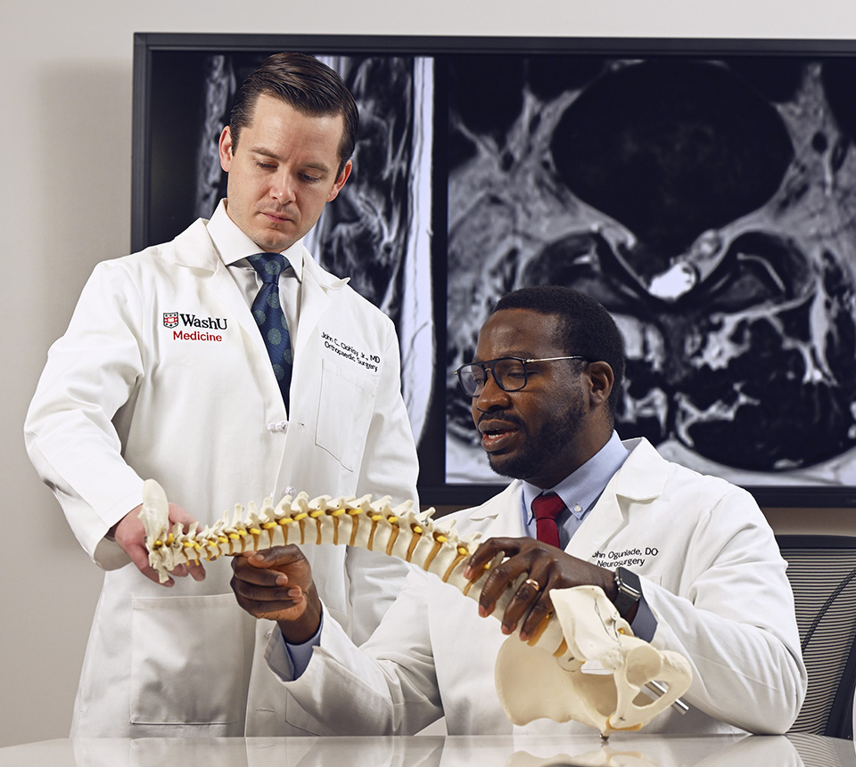 Two WashU Medicine surgeons examine a model of a human spine while sitting in front of MRI images of the spine.