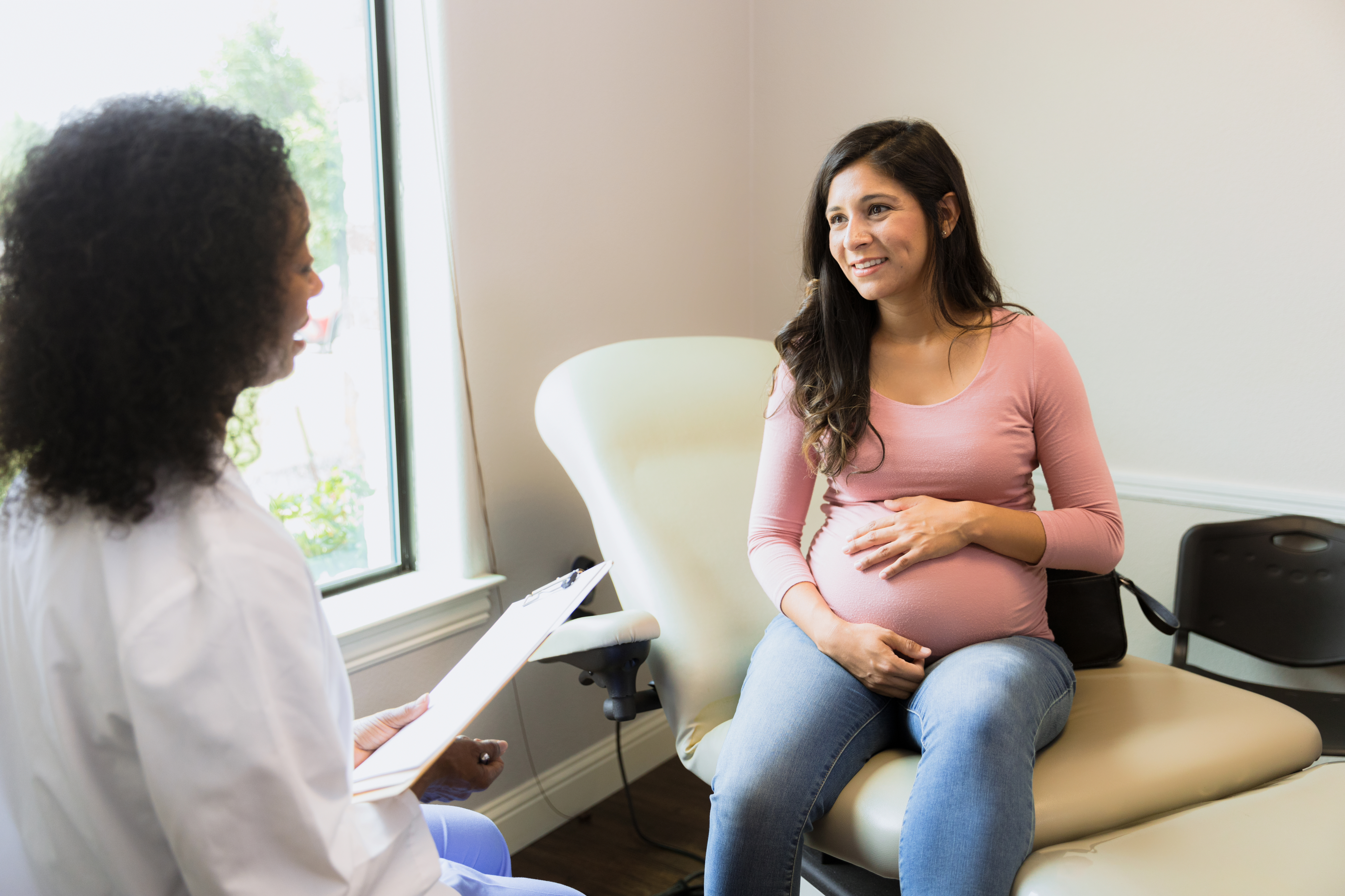 Pregnant woman with loose, curly black hair, pink top and jeans, sitting on hospital chair across from doctor in white coat