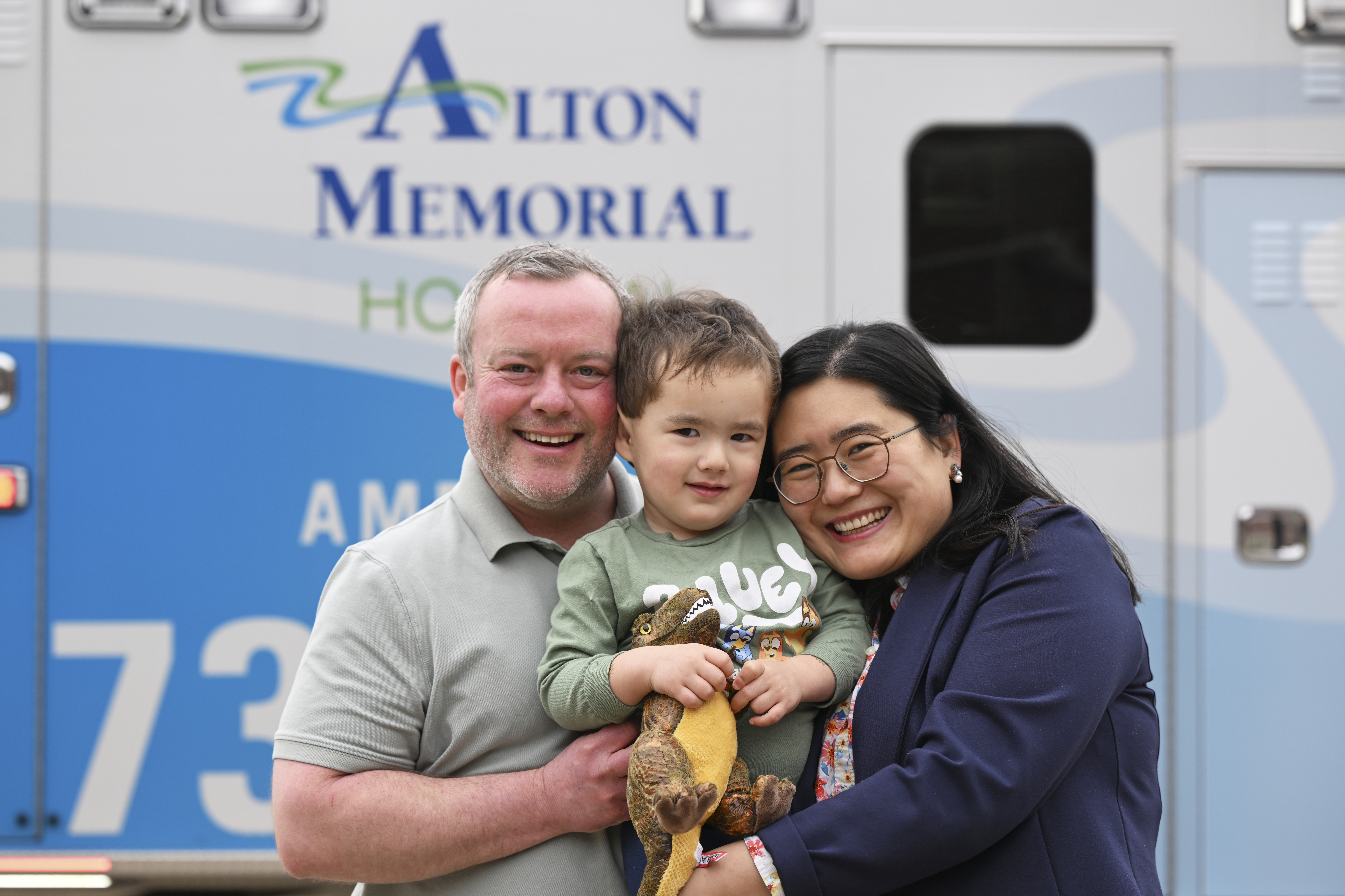 Half body shot of Ben (left) and Yaheng holding son in front of an Alton Memorial Hospital ambulance