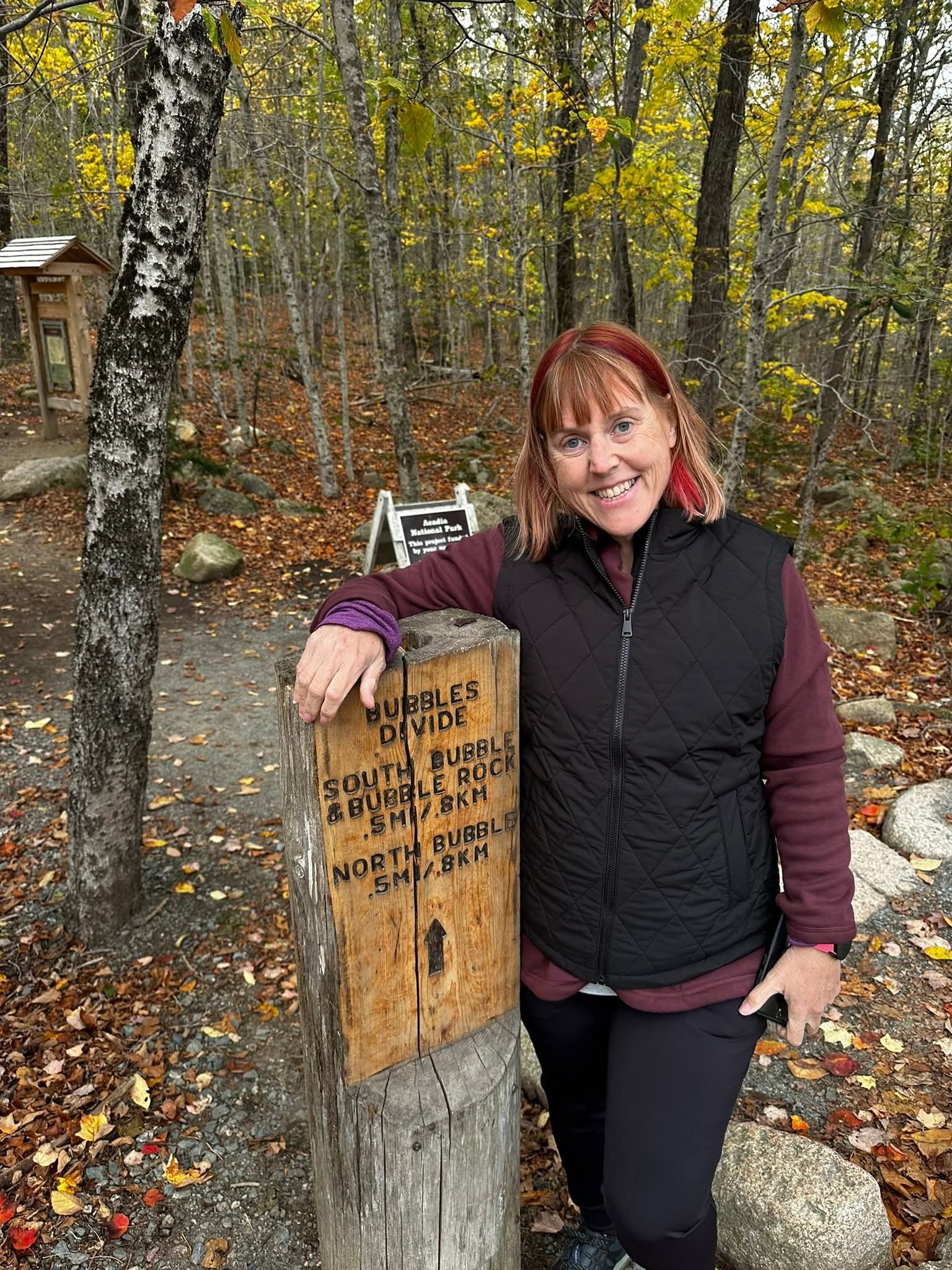 Patient Heather Taylor is smiling while standing next to a forest trail sign.