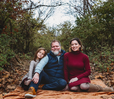 Abby (right) in a dark red sweaterdress, next to her husband and daughter in a forest background