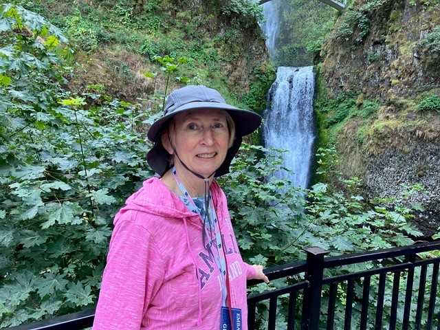 Charlotte Moro, 72, wearing pink thin jacket and black hat, standing in front of waterfall 