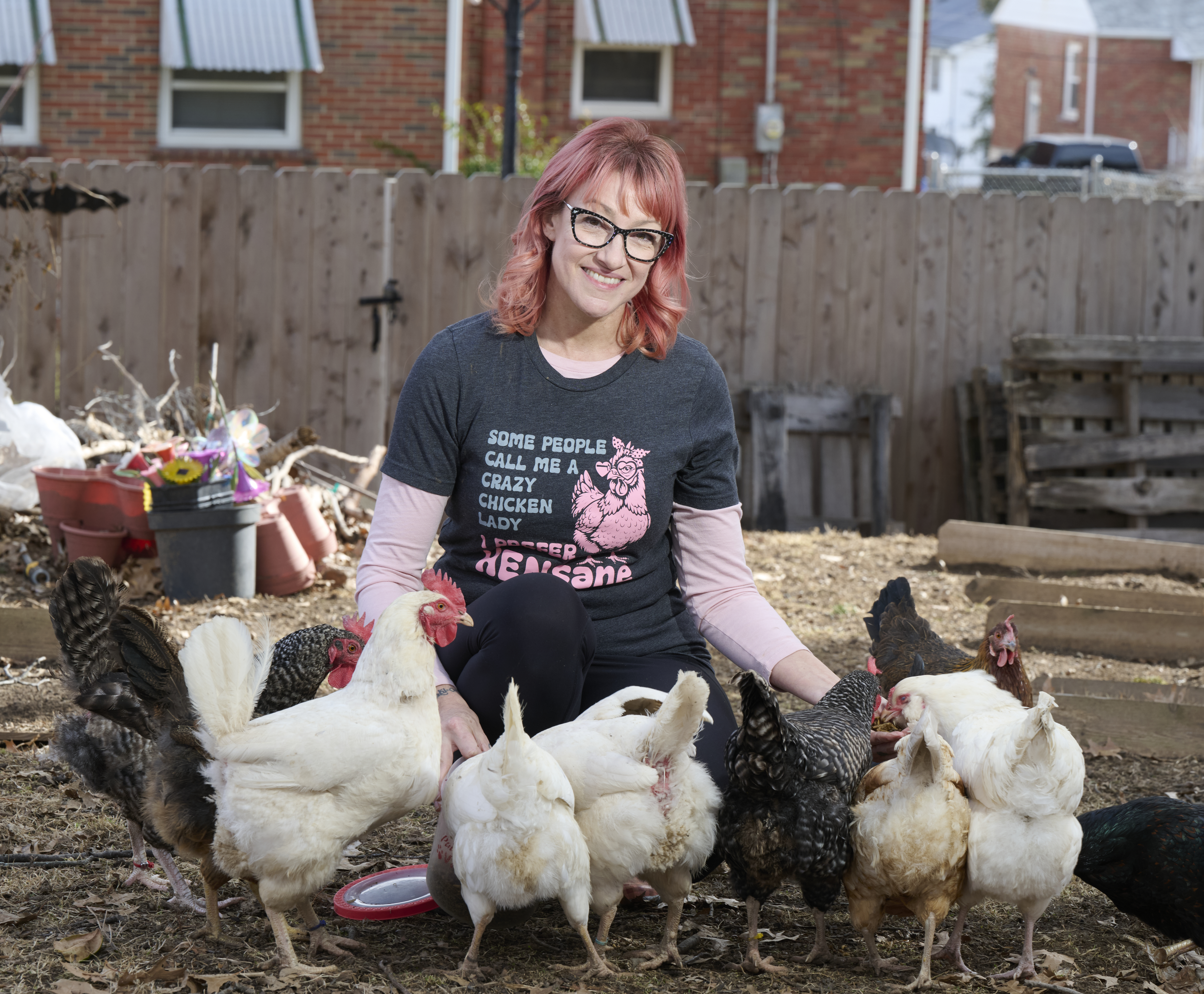 Debbi Jesse, 51, has red hair, glasses, and a black shirt with text, sitting with several chickens outdoors