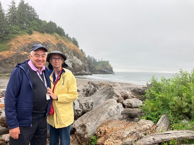 Joe (left) wears navy jacket and black hat, standing with wife Charlotte, 72 who wears yellow jacket and black hat, in front of a beach area