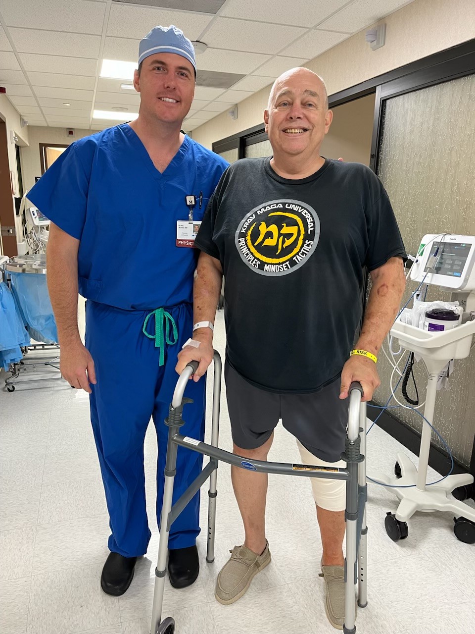 Dr. Busse (left) wears blue hospital scrubs, standing next to patient Kevin Sweeney, 71, who wears a black shirt and shorts and a walker. 