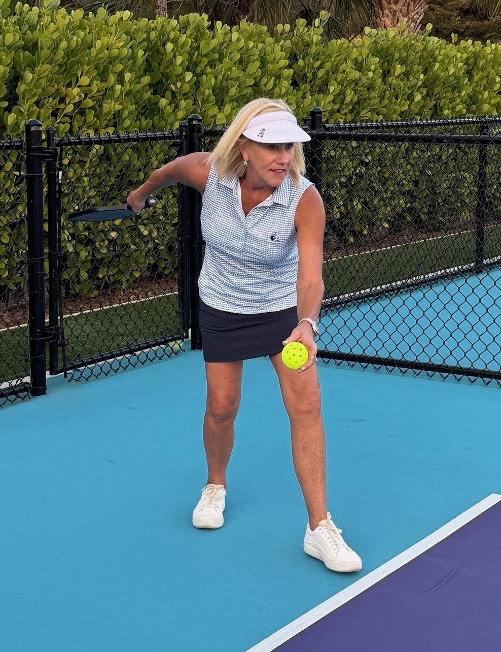 Barb Mitchell getting ready to serve the pickleball on a court, wearing a white cap and athletic top and skirt.