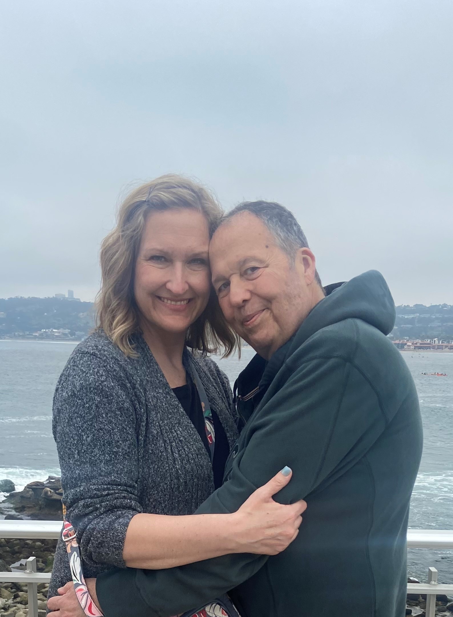 John Carney (left) and his wife wearing grey clothing, standing in a lakeside background