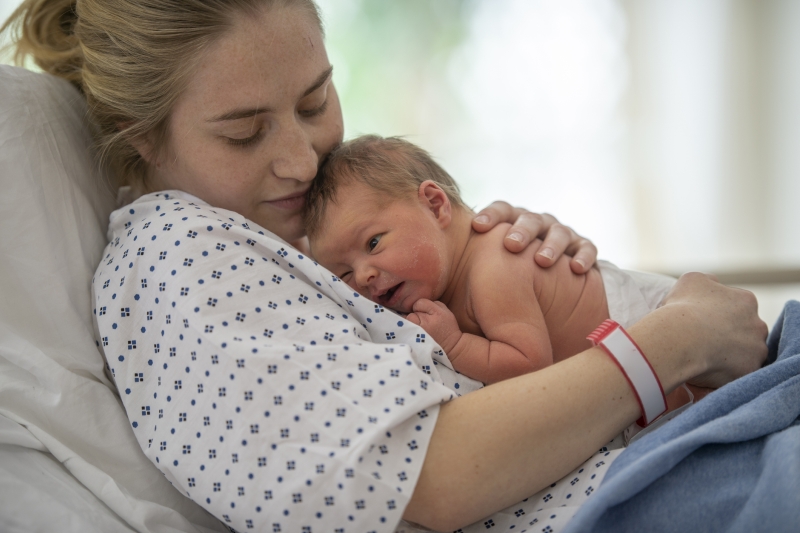 Woman with blonde hair tied back, wearing a hospital gown, lays on bed, holding a newborn baby